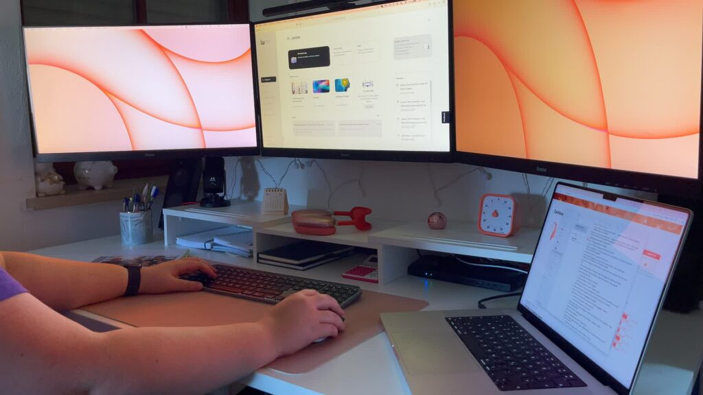 Ein Schreibtisch mit 3 Bildschirmen und zusätzlich rechts einem Macbook Laptop. Eine Person tippt auf einer Tastatur.